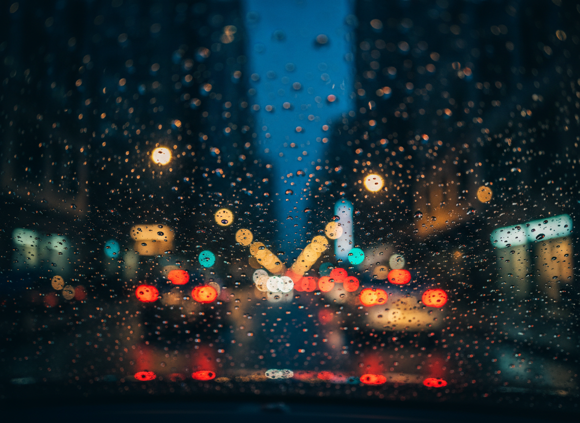 A moody nighttime view of a city under rain, seen through a car windshield covered in beaded droplets, rendered in rich photographic realism. Outside, blurred streetlights, traffic signals, and illuminated shopfronts dissolve into abstract patches of color, distorted by the water on the glass. The primary focus is on the sharply detailed raindrops in the foreground, each catching tiny reflections of the urban scene. Distant buildings form soft geometric silhouettes in cool blues and deep blacks. Oncoming headlights create subtle bokeh. The lighting is low and cinematic, with high contrast and selective focus creating a poetic, introspective atmosphere that feels like a frame from a film, embodying cinematic street photography without any visible figures.
